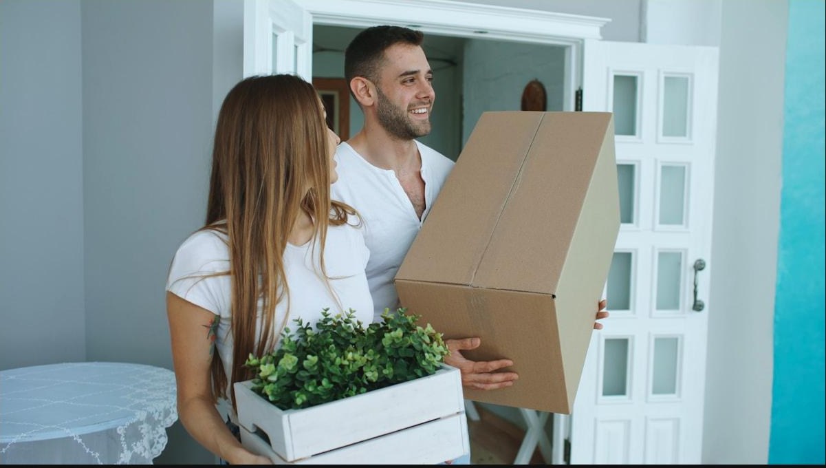 A couple carrying a box and a plant through the front door of their new home